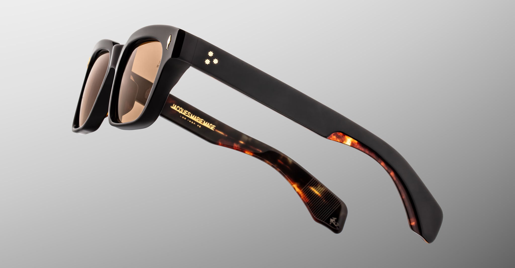 The Jacques Marie Mage Molino sunglasses by COMING SOON feature thick black frames, brown tinted lenses, tortoiseshell temples, and small white star details, displayed against a light gray background.
