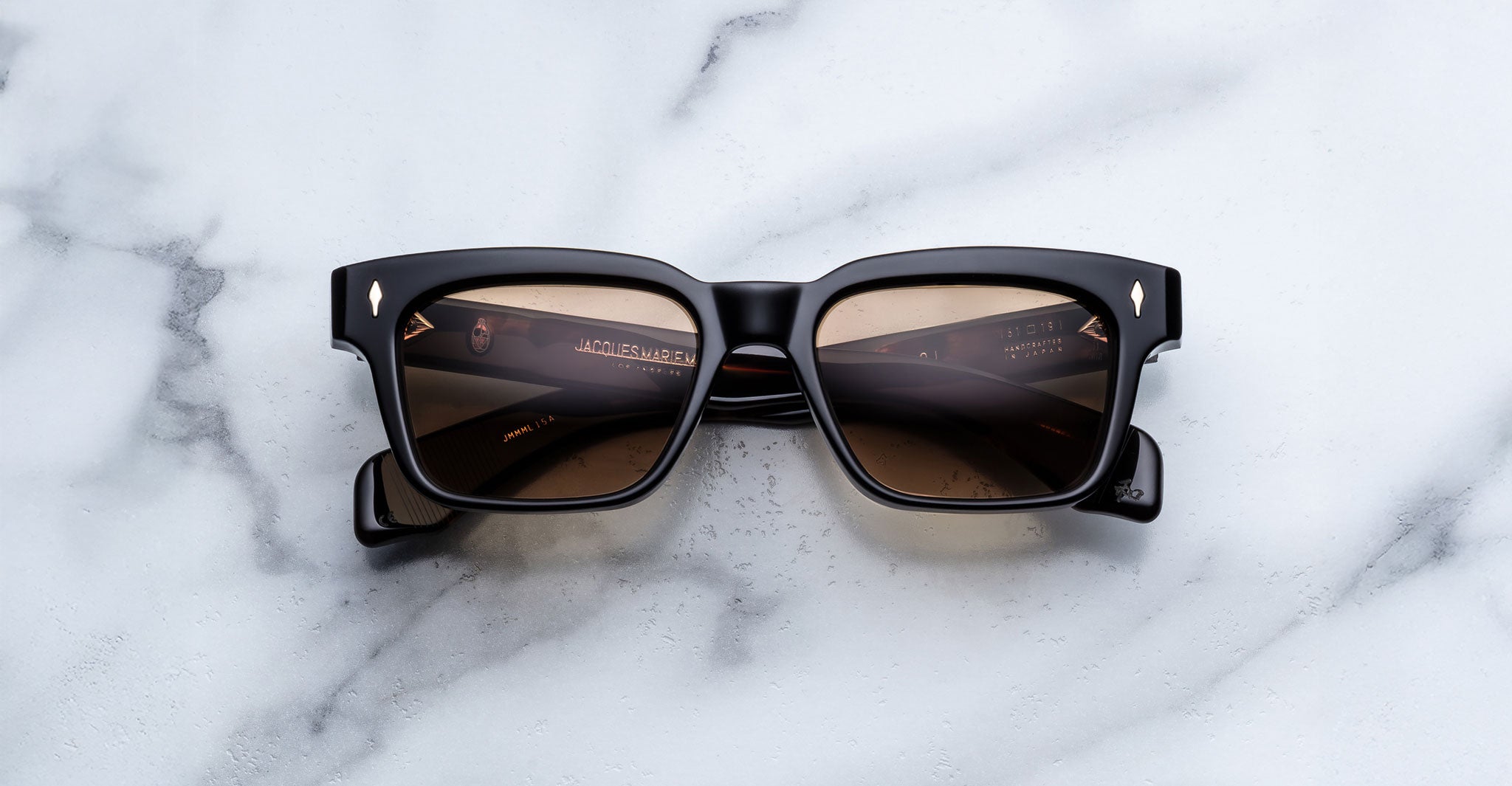 The Jacques Marie Mage Molino by COMING SOON, black square-framed sunglasses with brown-tinted lenses and gold-accented arms, rests on a white marble surface; subtle branding appears inside the temples.