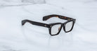 The Jacques Marie Mage Molino eyeglasses by COMING SOON feature bold, black rectangular frames and are displayed on a white marble surface.