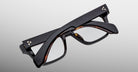 A pair of Jacques Marie Mage Molino black eyeglasses by COMING SOON, folded flat with arms crossed, displayed on a plain light gray background.