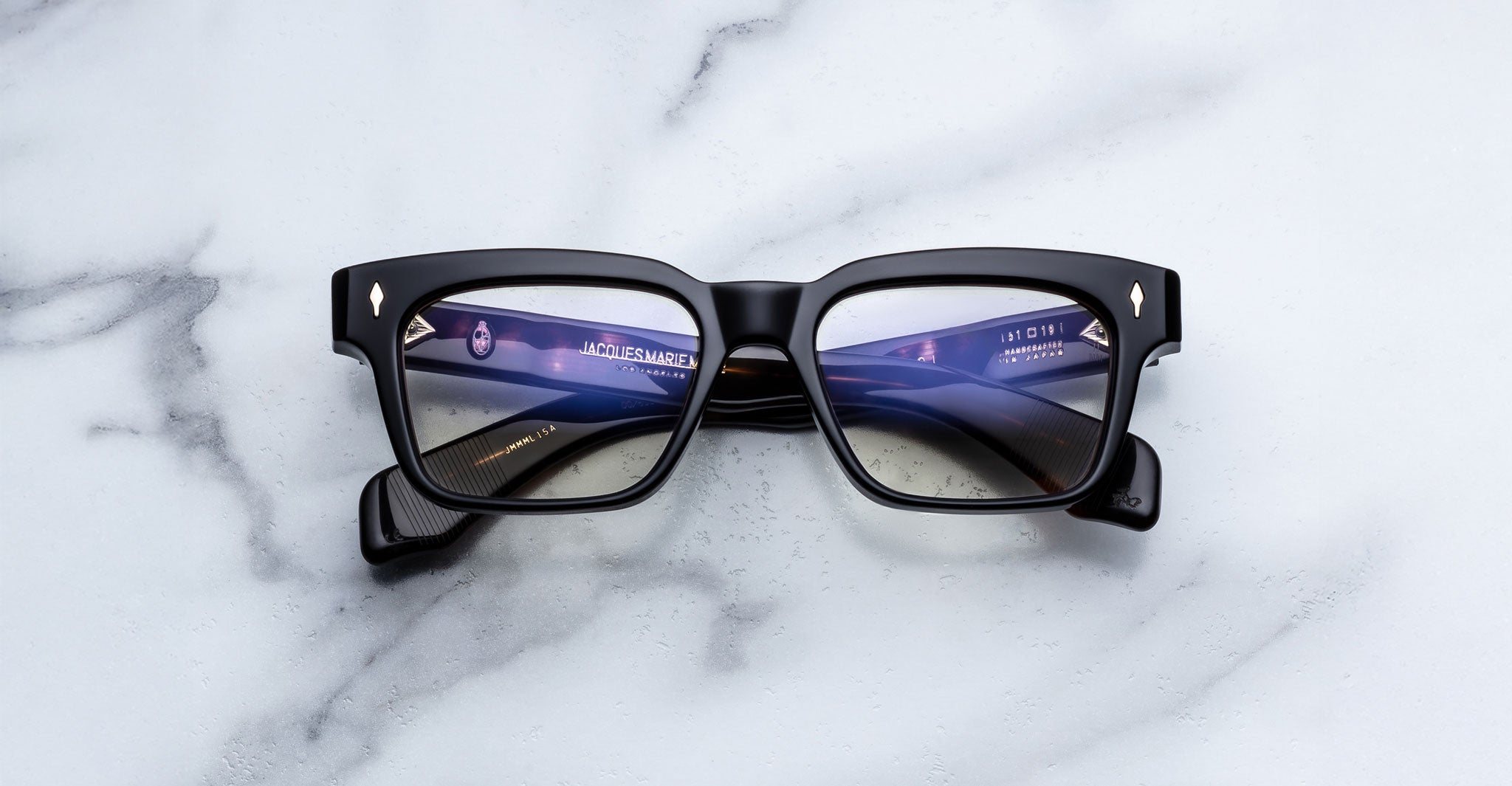 The Jacques Marie Mage Molino by COMING SOON features black square frames and blue-tinted lenses, shown resting on a white marble surface with screen reflections visible on the lenses.