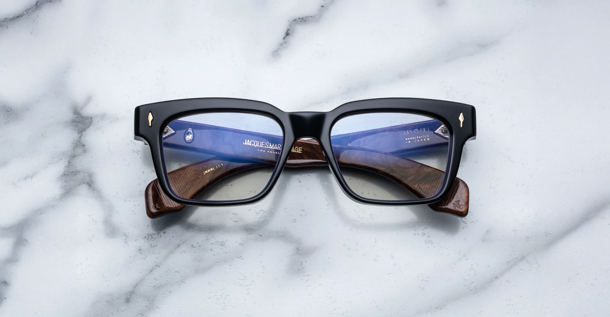 The Jacques Marie Mage Molino is a pair of rectangular eyeglasses with black frames and brown temples, displayed on a white marble surface. Reflections in the lenses reveal text and markings inside the arms.