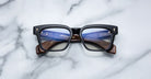 The Jacques Marie Mage Molino is a pair of rectangular eyeglasses with black frames and brown temples, displayed on a white marble surface. Reflections in the lenses reveal text and markings inside the arms.