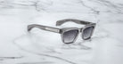 The Jacques Marie Mage Molino by COMING SOON, featuring silver frames and dark lenses, rests on a white marble surface with gray veining.