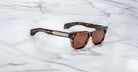 The Jacques Marie Mage Molino are tortoiseshell sunglasses with rectangular brown lenses, wide arms, and a metallic accent by the hinges, displayed on a white marble surface.