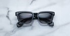 A pair of Jacques Marie Mage Molino black square-framed sunglasses with dark lenses is displayed on a white marble surface.