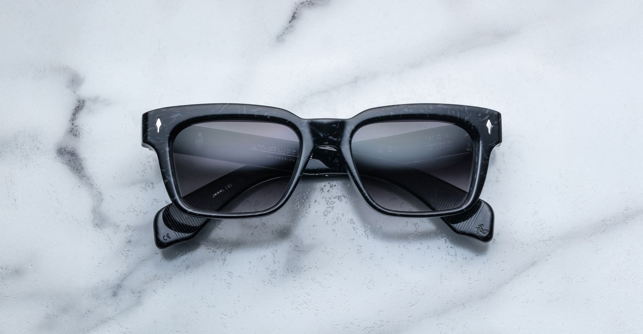 A pair of Jacques Marie Mage Molino black square-framed sunglasses with dark lenses is displayed on a white marble surface.