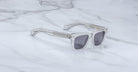 The Jacques Marie Mage Molino sunglasses by COMING SOON, featuring clear frames and dark lenses, rest on a white marble surface.