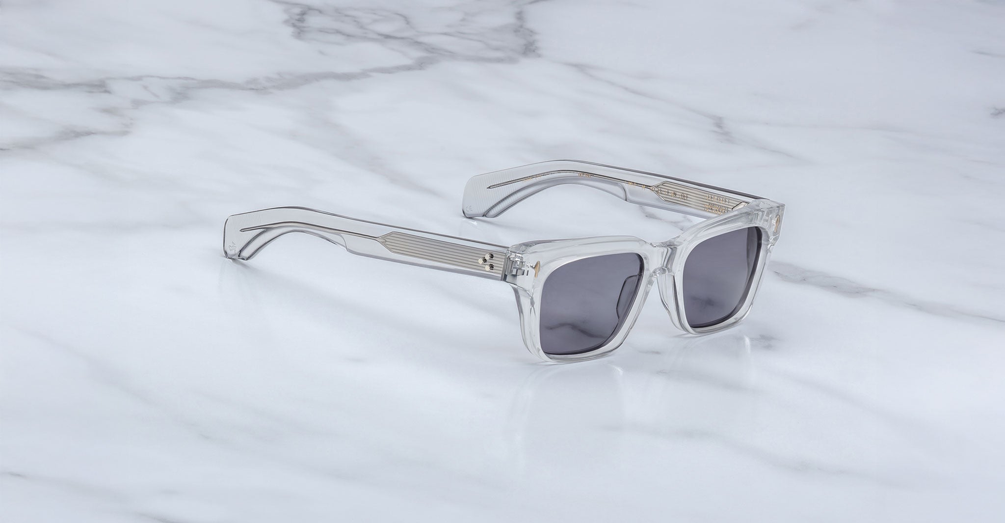 The Jacques Marie Mage Molino sunglasses by COMING SOON, featuring clear frames and dark lenses, rest on a white marble surface.