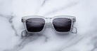 A pair of Jacques Marie Mage Molino square, translucent gray sunglasses by COMING SOON with dark lenses rests on white marble, reflecting a ceiling light pattern.