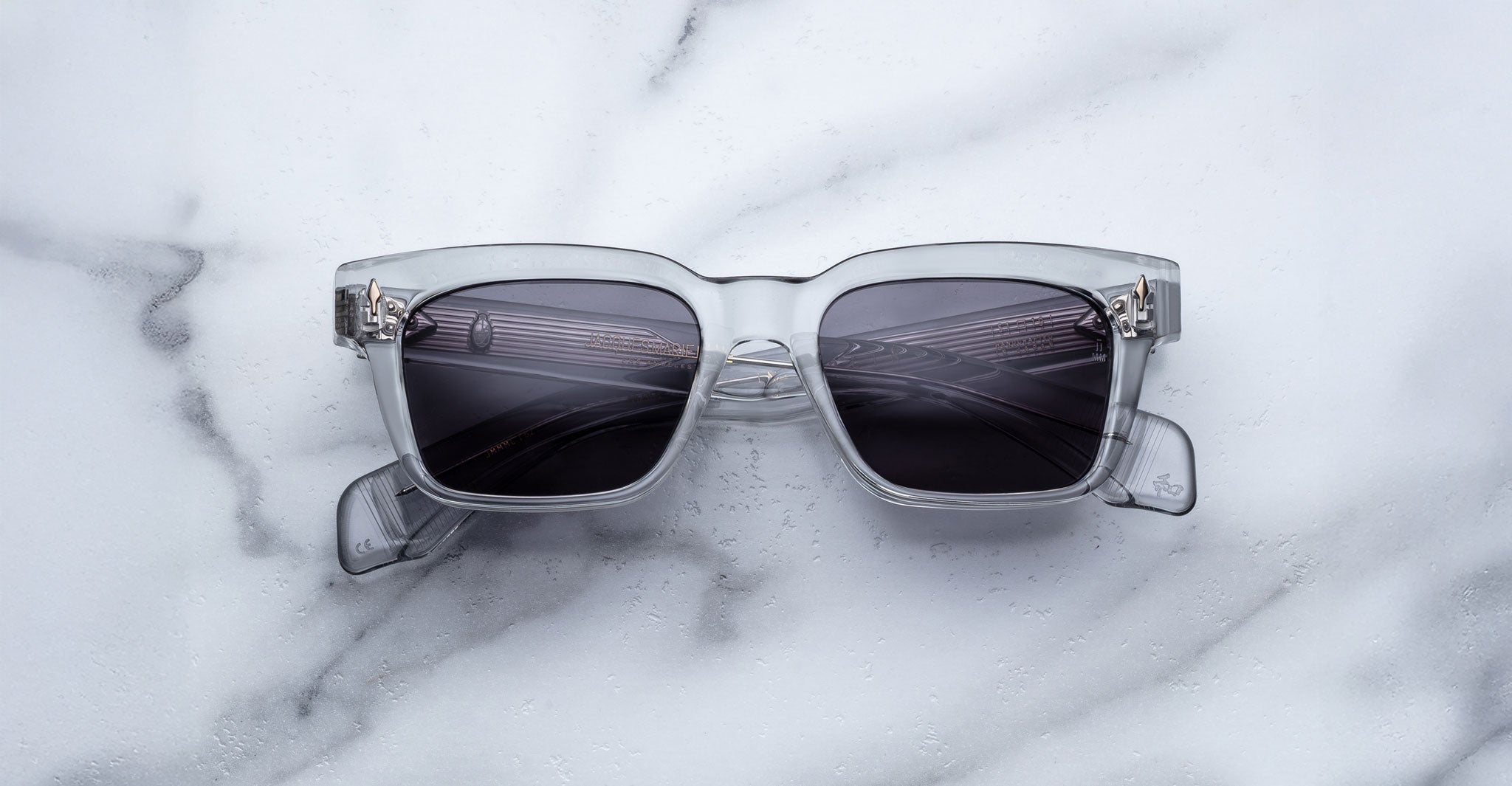 A pair of Jacques Marie Mage Molino square, translucent gray sunglasses by COMING SOON with dark lenses rests on white marble, reflecting a ceiling light pattern.