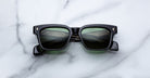 A pair of Jacques Marie Mage Molino black rectangular sunglasses with dark lenses and small gold accents near the hinges rests on a white marble surface.