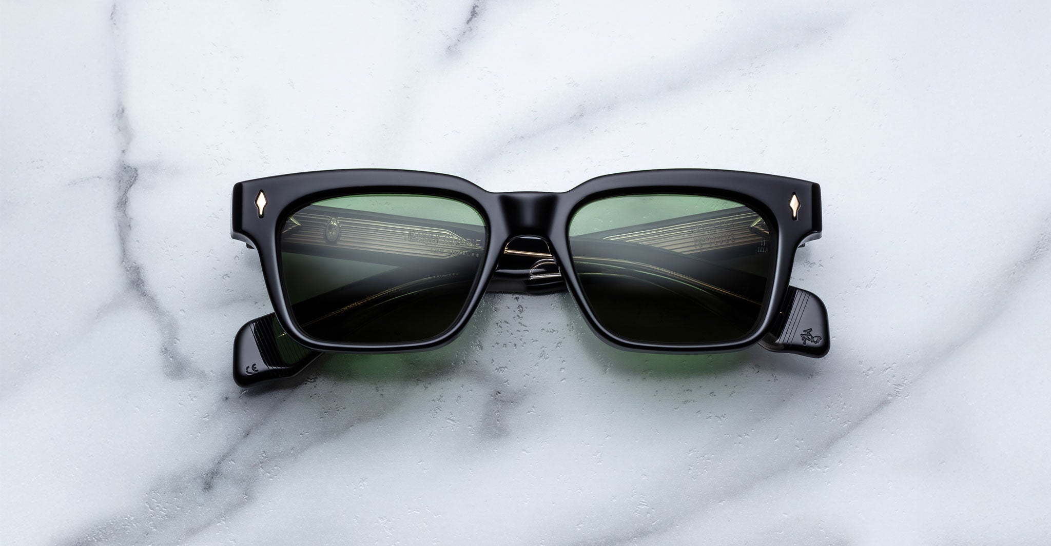 A pair of Jacques Marie Mage Molino black rectangular sunglasses with dark lenses and small gold accents near the hinges rests on a white marble surface.