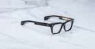 The Jacques Marie Mage Molino featuring black rectangular thick frames and clear lenses, rests on a white marble surface.