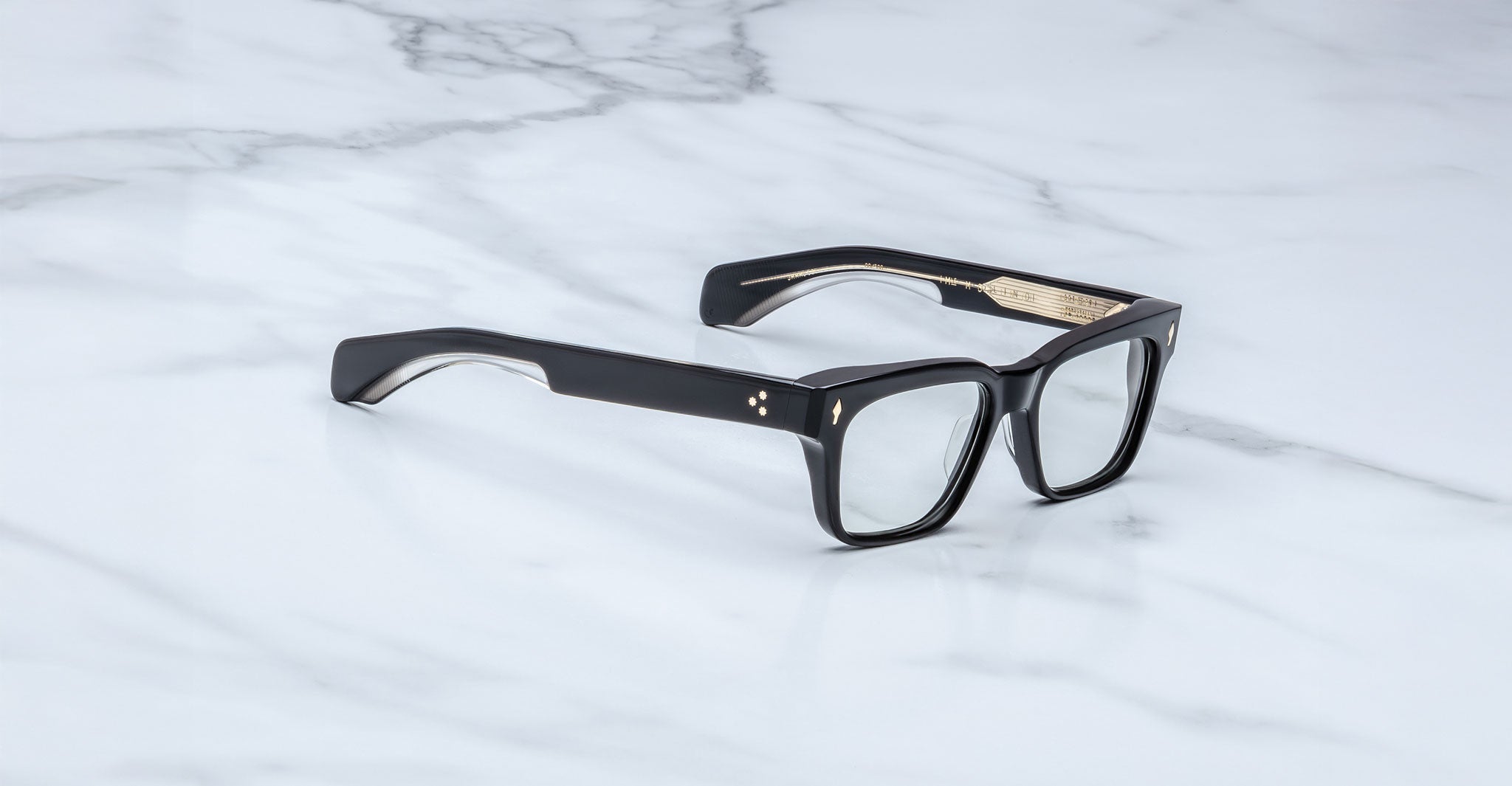 The Jacques Marie Mage Molino featuring black rectangular thick frames and clear lenses, rests on a white marble surface.