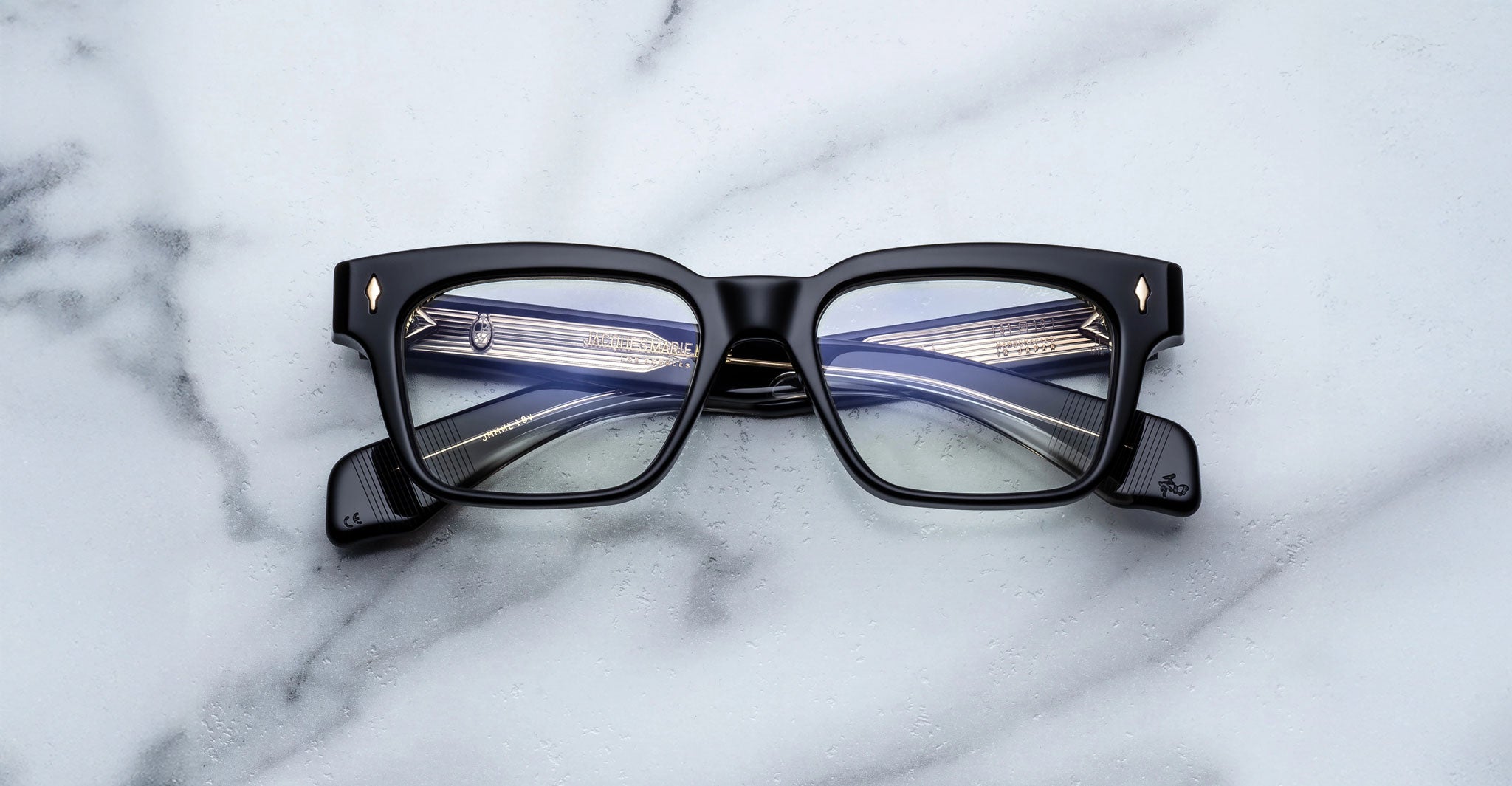 The Jacques Marie Mage Molino by COMING SOON, black rectangular eyeglasses with thick frames, rest on a white marble surface, their lenses reflecting the temples and hinges.