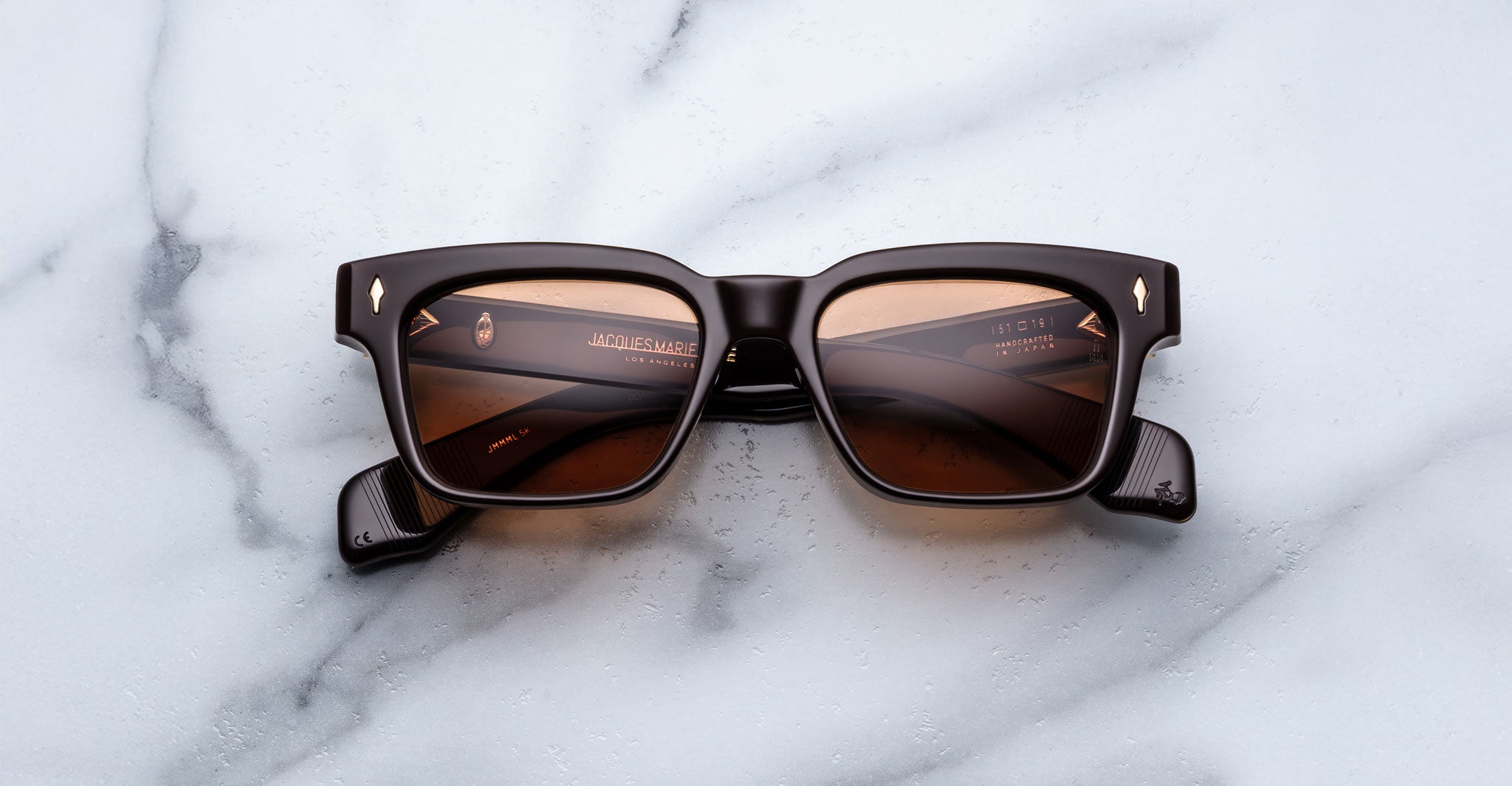 A pair of Jacques Marie Mage Molino square sunglasses with black frames and brown-tinted lenses is displayed face-up on a white marble surface.
