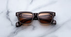A pair of Jacques Marie Mage Molino square sunglasses with black frames and brown-tinted lenses is displayed face-up on a white marble surface.
