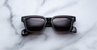 Jacques Marie Mage Molino black rectangular sunglasses with dark tinted lenses and gold diamond-shaped accents on the upper corners, displayed on a white marble surface.
