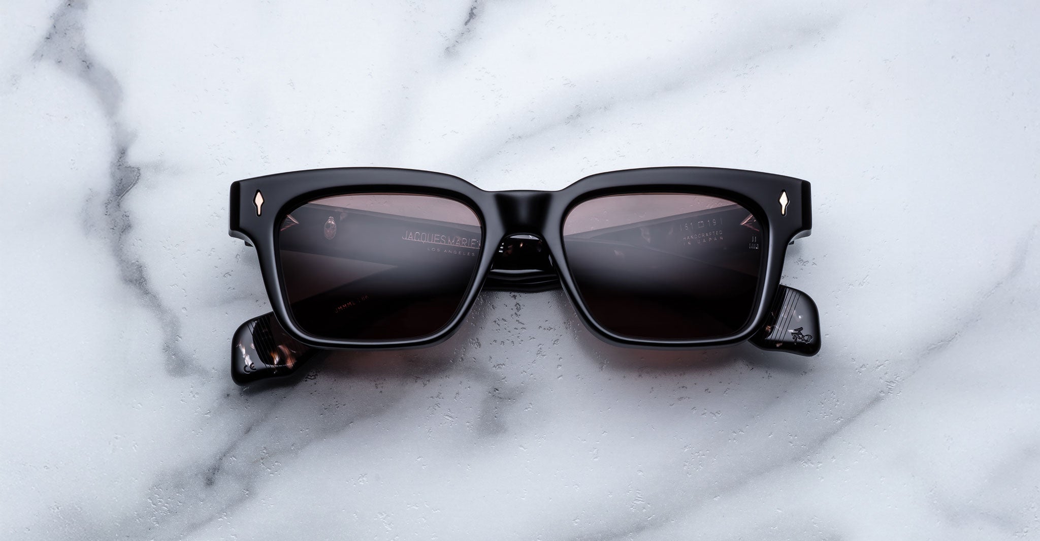 Jacques Marie Mage Molino black rectangular sunglasses with dark tinted lenses and gold diamond-shaped accents on the upper corners, displayed on a white marble surface.