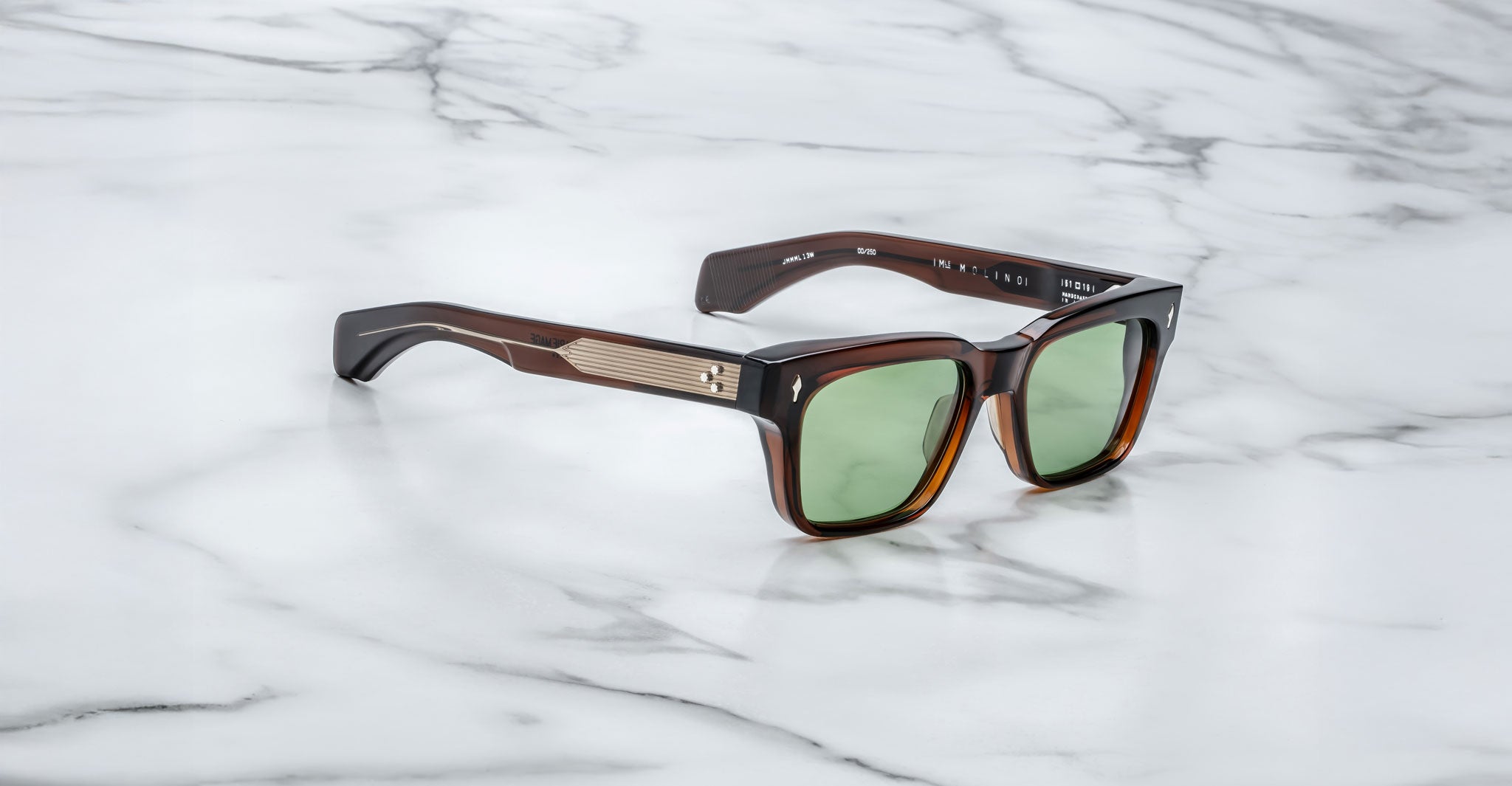 The Jacques Marie Mage Molino sunglasses feature a stylish square brown frame with green lenses and metallic temple accents, displayed on a white marble surface.