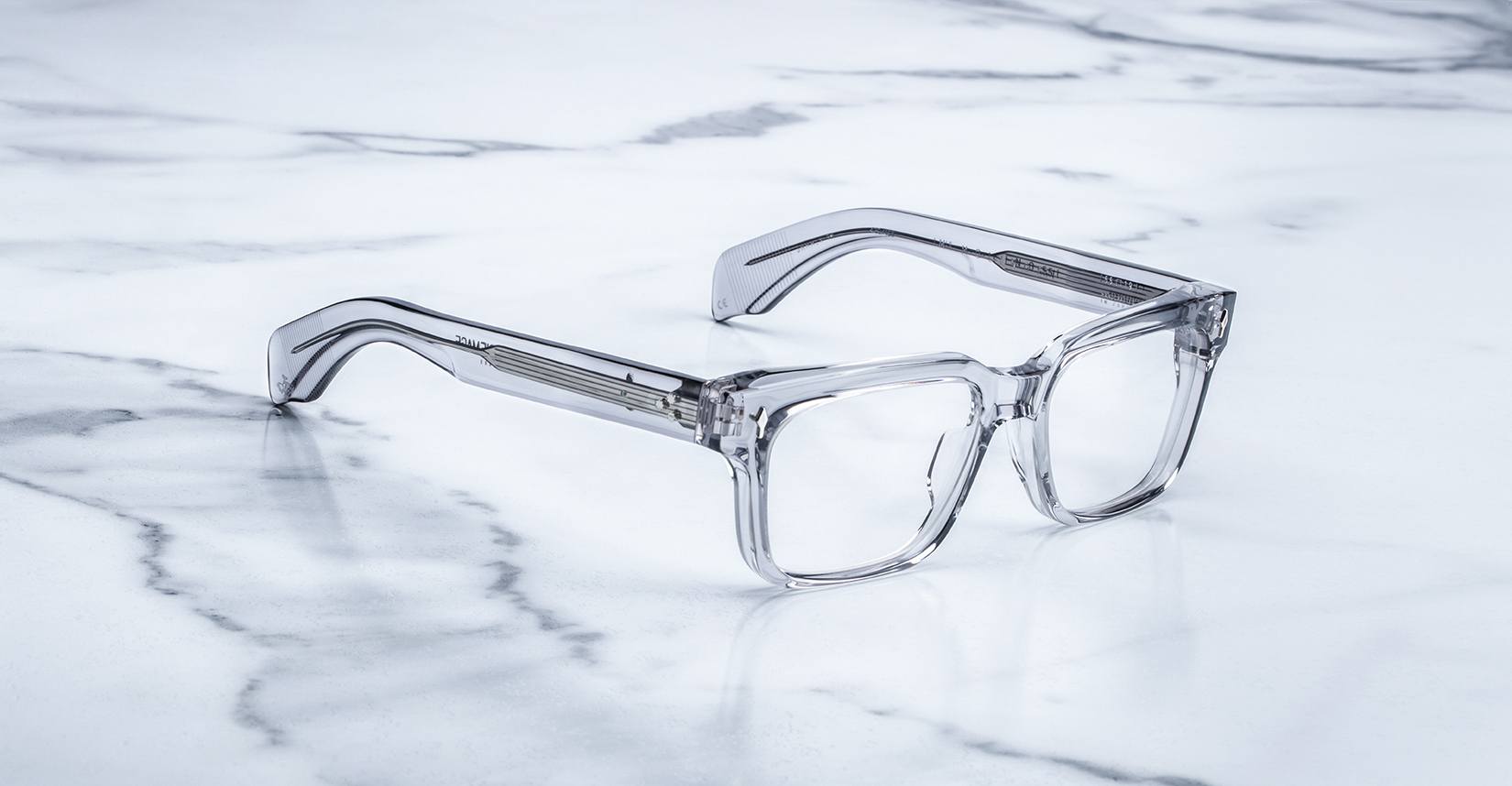 The Jacques Marie Mage Molino 55 features clear, rectangular eyeglasses with thick transparent frames, showcased on a white marble surface with subtle gray veining.