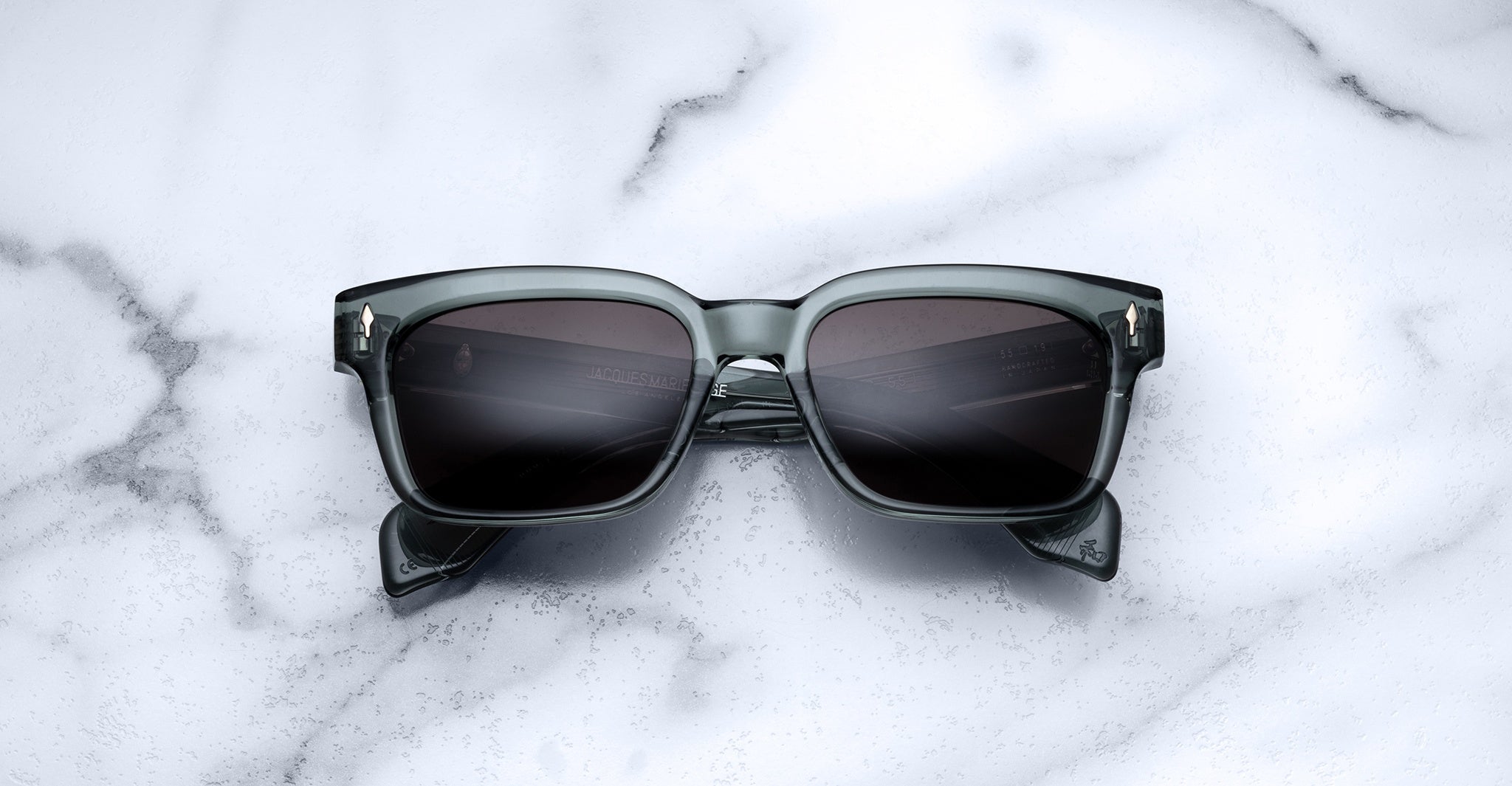A pair of Jacques Marie Mage Molino 55 square-framed sunglasses with dark lenses rests on a white marble surface.