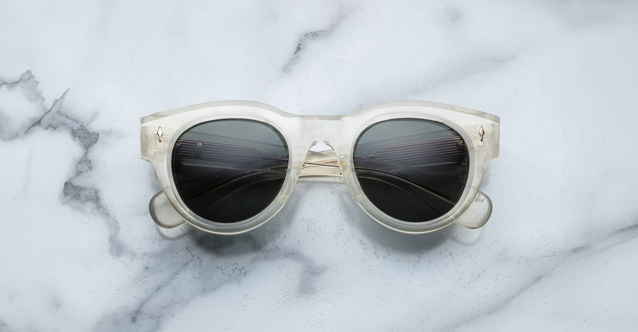 Jacques Marie Mage Molitor sunglasses with clear frames and dark lenses displayed on a white marble surface with grey veining.