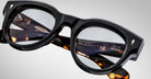 Close-up of Jacques Marie Mage Molitor eyeglasses featuring round black and tortoiseshell frames. Brand markings and product details are visible on the inside of the temples. The frames rest on a white surface with a gradient background.