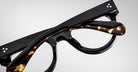 A close-up of folded Jacques Marie Mage Molitor eyeglasses with black and tortoiseshell-patterned frames, resting on a light gray background.