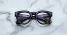 The Jacques Marie Mage Molitor black eyeglasses feature round lenses and tortoiseshell-patterned temple tips, resting on a white marble surface. The arms and text can be seen through the lenses.