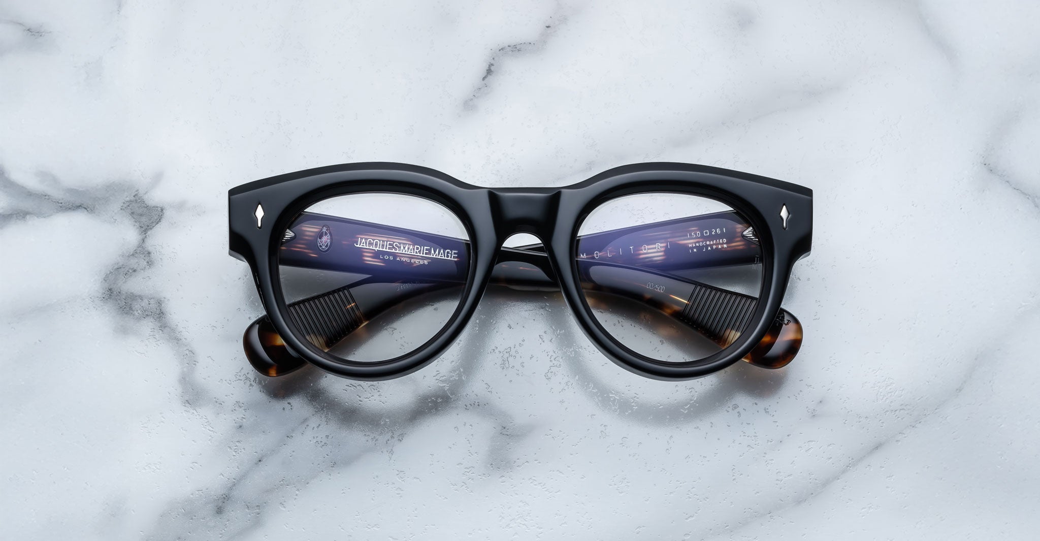 The Jacques Marie Mage Molitor black eyeglasses feature round lenses and tortoiseshell-patterned temple tips, resting on a white marble surface. The arms and text can be seen through the lenses.
