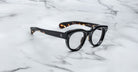 A pair of Jacques Marie Mage Molitor eyeglasses with black thick rims, rounded frames, and tortoiseshell-patterned temples rests on a white marble surface with grey veining.