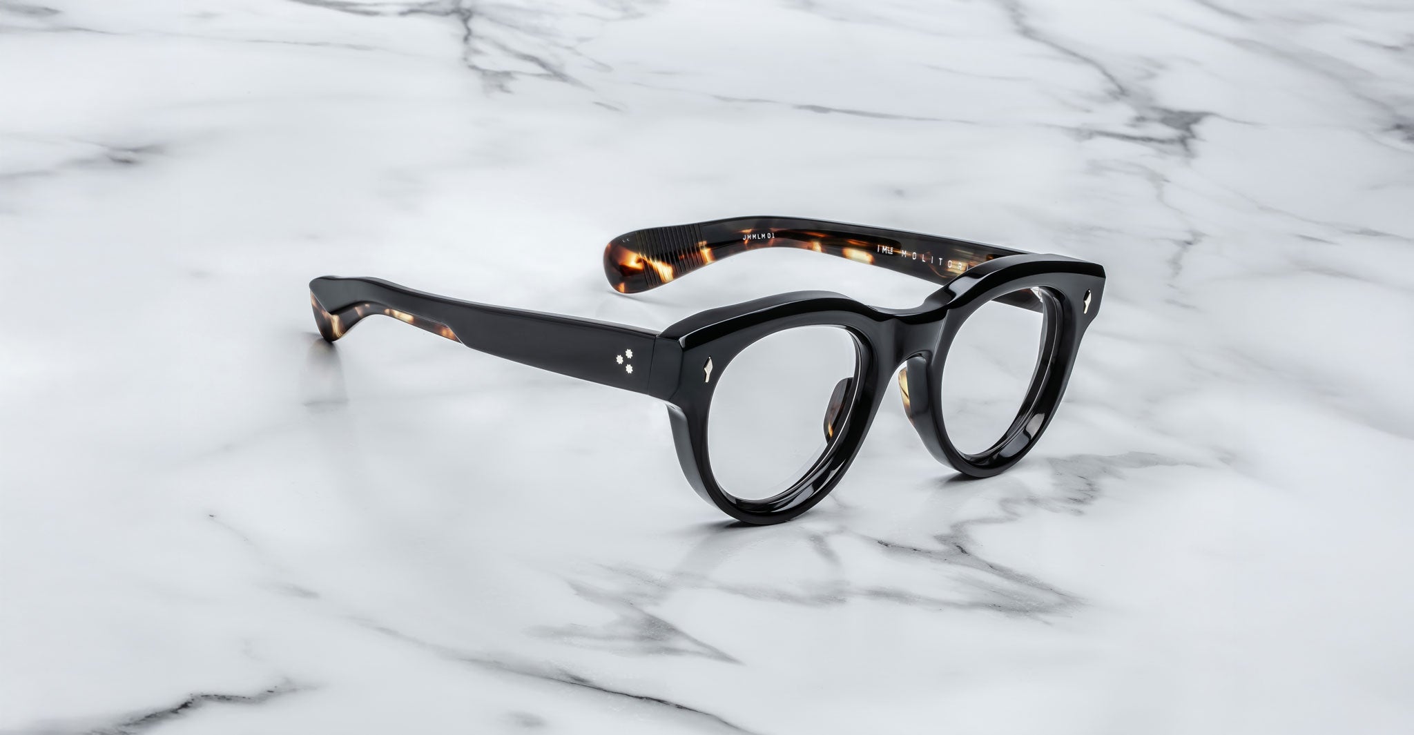 A pair of Jacques Marie Mage Molitor eyeglasses with black thick rims, rounded frames, and tortoiseshell-patterned temples rests on a white marble surface with grey veining.