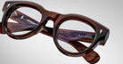 Close-up of Jacques Marie Mage Molitor eyeglasses—thick, glossy brown frames with clear lenses, featuring engraved brand and model info on the inner temple arms, set against a plain light background.