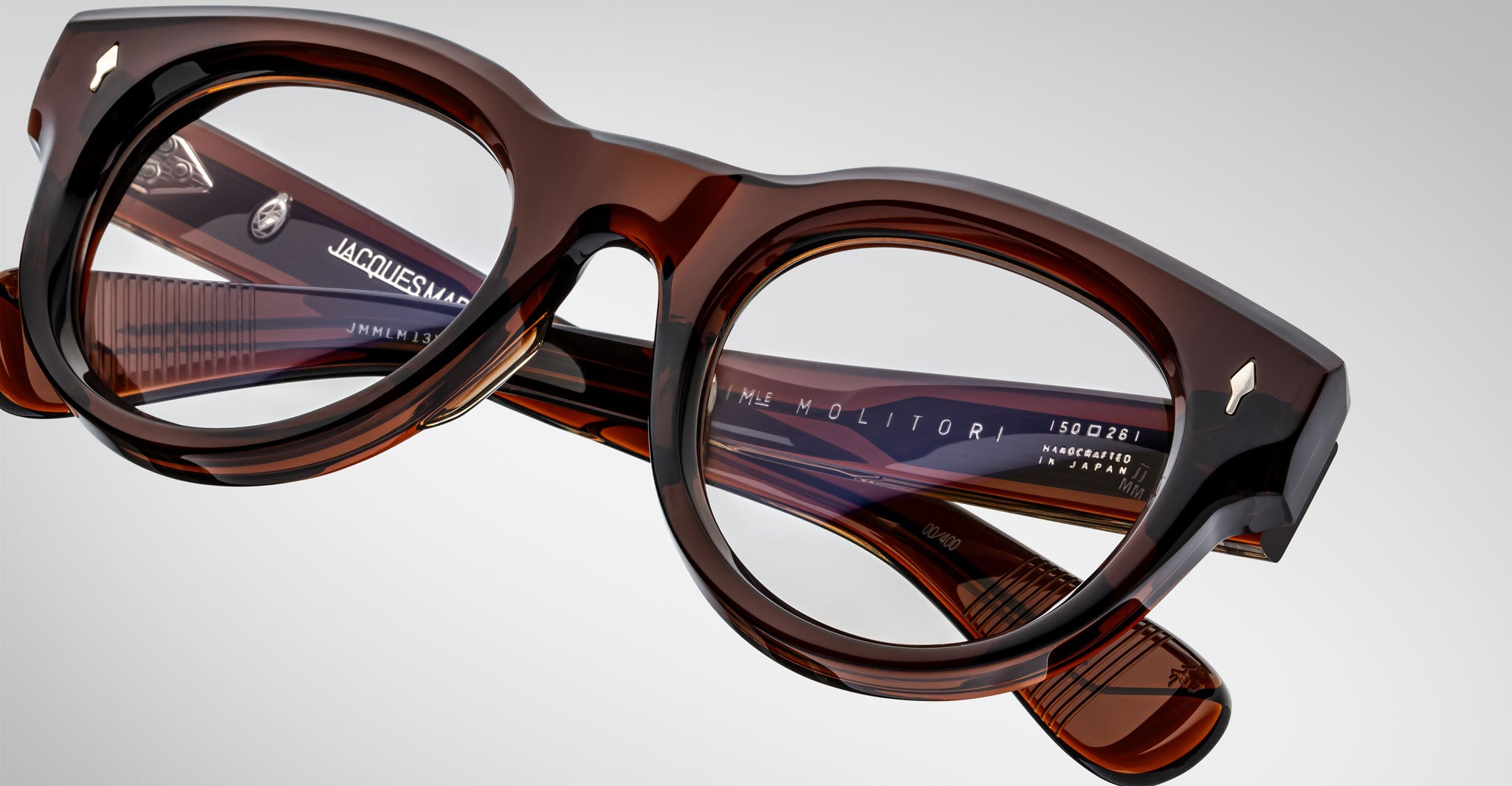 Close-up of Jacques Marie Mage Molitor eyeglasses—thick, glossy brown frames with clear lenses, featuring engraved brand and model info on the inner temple arms, set against a plain light background.