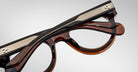 A close-up of the Jacques Marie Mage Molitor eyeglasses in brown and black with silver detailing on the temples, shown folded on a plain light background.