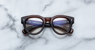 The Jacques Marie Mage Molitor features thick, round, dark brown frames with silver accents, displayed on a white marble surface. Branding and text are visible on the inside of the arms.