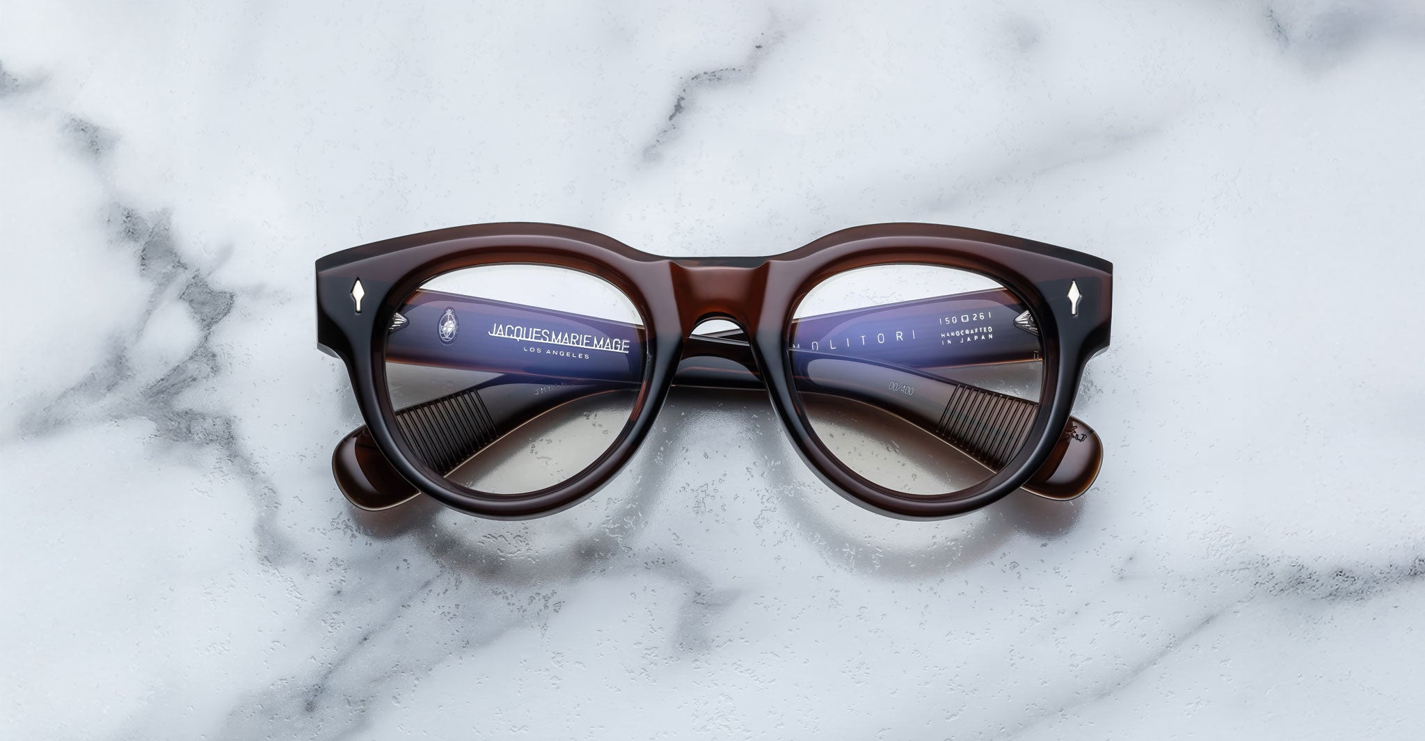 The Jacques Marie Mage Molitor features thick, round, dark brown frames with silver accents, displayed on a white marble surface. Branding and text are visible on the inside of the arms.