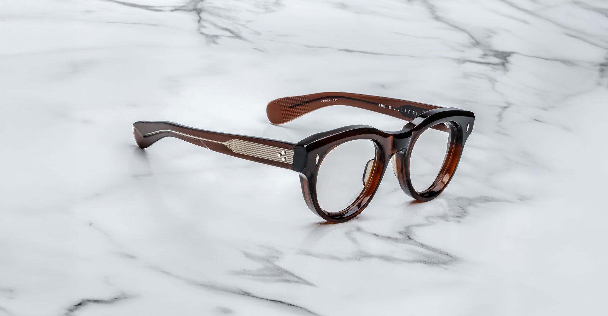 A pair of Jacques Marie Mage Molitor brown, thick-framed eyeglasses with metallic temple accents rests on a white marble surface with gray veining.