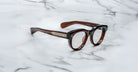 A pair of Jacques Marie Mage Molitor brown, thick-framed eyeglasses with metallic temple accents rests on a white marble surface with gray veining.