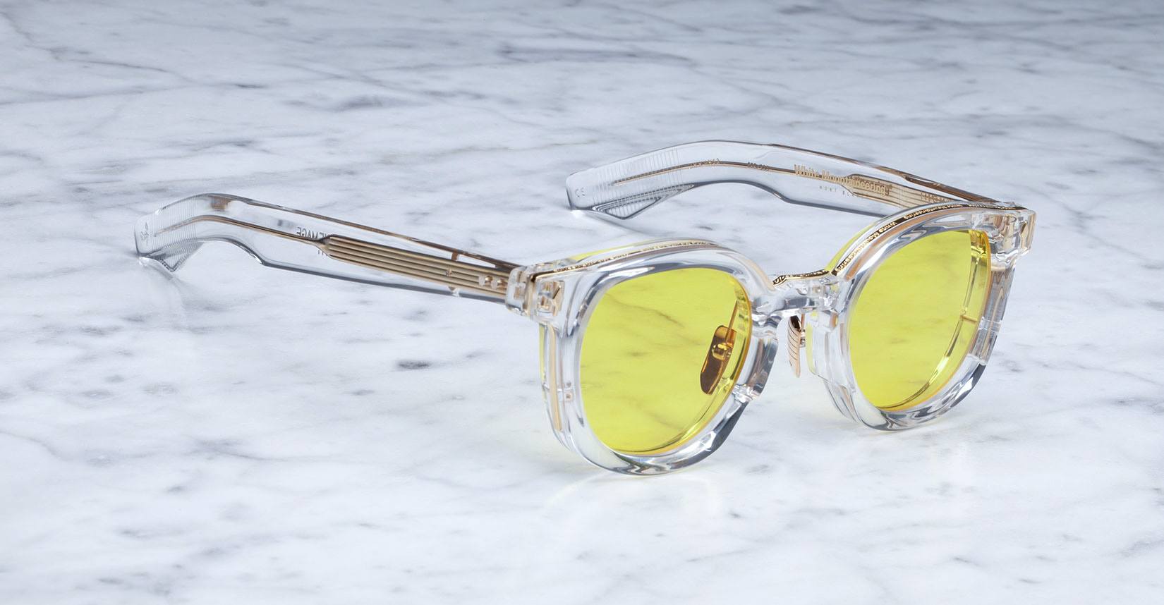 The Jacques Marie Mage Mont Blanc eyeglasses, featuring clear frames with yellow-tinted lenses and a transparent glossy finish on the temples and bridge, rest on a light gray marble surface.