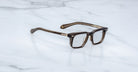 Jacques Marie Mage Moscova in London acetate optical frame, angled view with rectangular lenses, translucent brown acetate front, signature arrowhead pins, and exposed core wire temples with warm tonal detailing