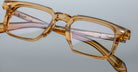 Close-up of Jacques Marie Mage Moscova eyeglasses, featuring thick rectangular translucent amber frames with visible hinges, engraved temples, and metal details, set against a plain gray background.