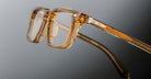 Close-up of Jacques Marie Mage Moscova eyeglasses featuring thick, transparent amber-colored frames and metal hinges, photographed against a gradient gray background.