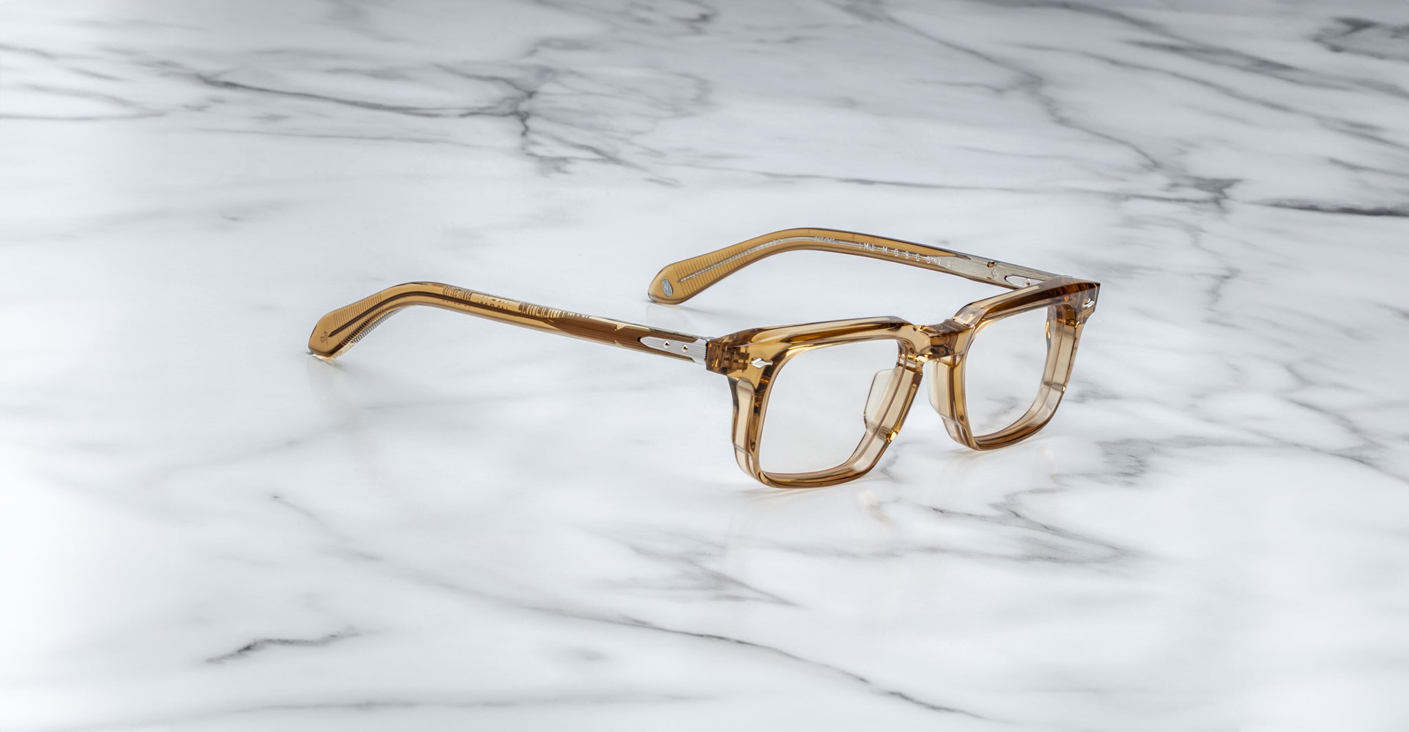 The Jacques Marie Mage Moscova brown transparent square-frame eyeglasses are displayed on a white marble surface with gray veining.