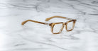 The Jacques Marie Mage Moscova brown transparent square-frame eyeglasses are displayed on a white marble surface with gray veining.