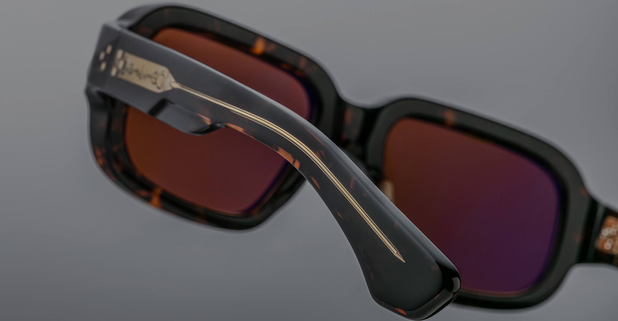 Close-up of Jacques Marie Mage Nakahira sunglasses: dark, rectangular frames with a subtle tortoiseshell pattern, tinted lenses, and a metallic engraved logo on the temple, set against a gray background.