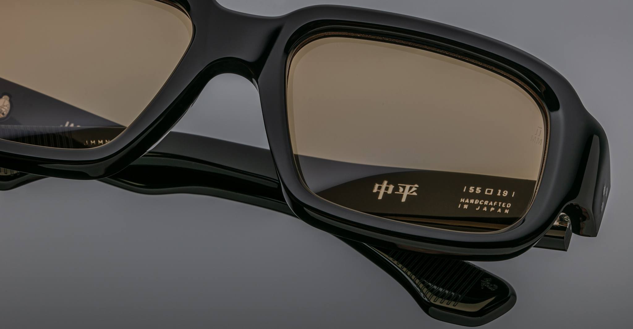 Close-up of Jacques Marie Mage Nakahira black eyeglasses with tan lenses. Handcrafted in Japan and Asian characters are visible on the inner temple against a smooth gray background.