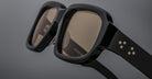 Close-up of Jacques Marie Mage Nakahira black rectangular sunglasses with gold star-shaped accents on the arms, set against a neutral gray background.